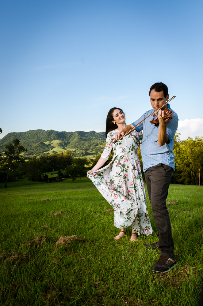 Foto do ensaio pre wedding, casamento, do casal ao ar livre com sol e céu azul tocando violino no Hotel Capo Zorial com piscina e monte ao fundo no Recanto do Maestro em São João do Polêsine, RS, próximo de Santa Maria, RS