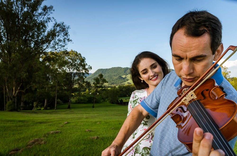 Foto do ensaio pre wedding, casamento, do casal ao ar livre com sol e céu azul tocando violino no Hotel Capo Zorial com piscina e monte ao fundo no Recanto do Maestro em São João do Polêsine, RS, próximo de Santa Maria, RS