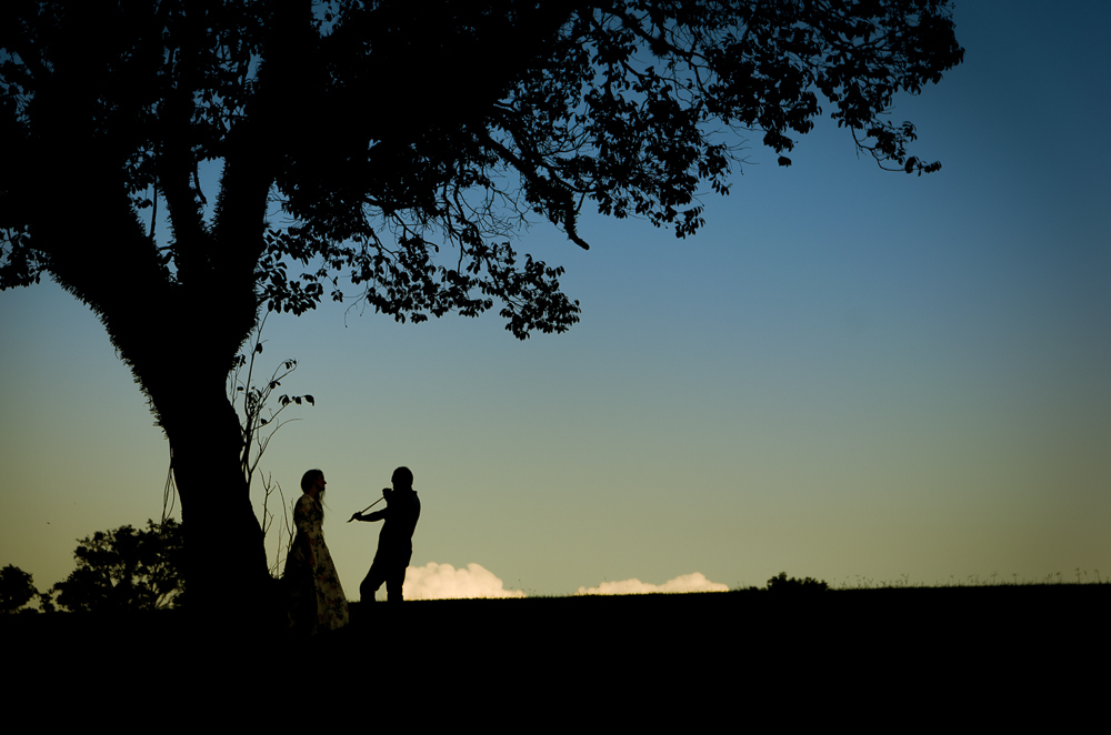 Foto do ensaio pre wedding, casamento, do casal ao ar livre com sol e céu azul tocando violino no Hotel Capo Zorial com piscina e monte ao fundo no Recanto do Maestro em São João do Polêsine, RS, próximo de Santa Maria, RS