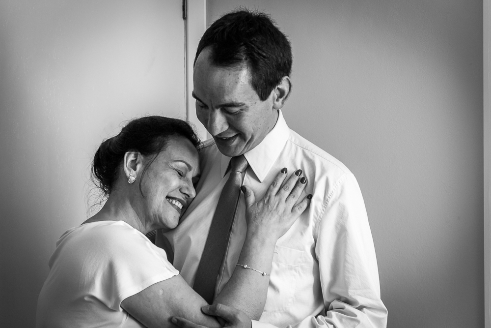 Foto do ensaio de making of do noivo no quarto fazendo os preparativos para o casamento. A mãe do noivo o abraça com ternura no quarto em Restinga Seca, RS 