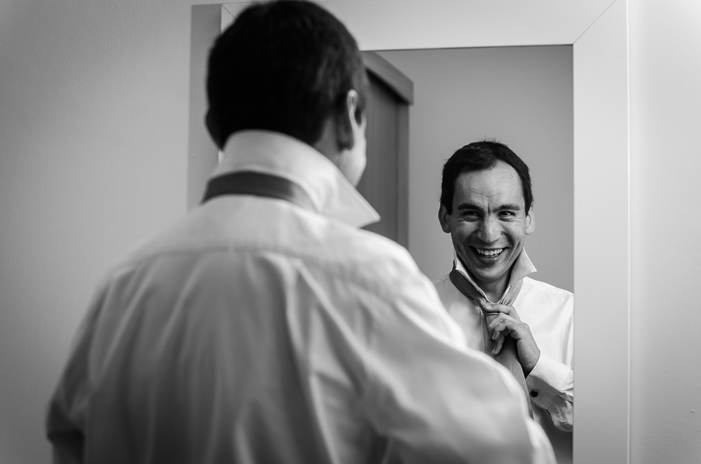 Foto  em preto e brando, PB, do ensaio de making of do noivo no quarto fazendo os preparativos para o casamento. Está dando o nó na gravata se olhando e sorrindo no espelho em Restinga Seca, RS 