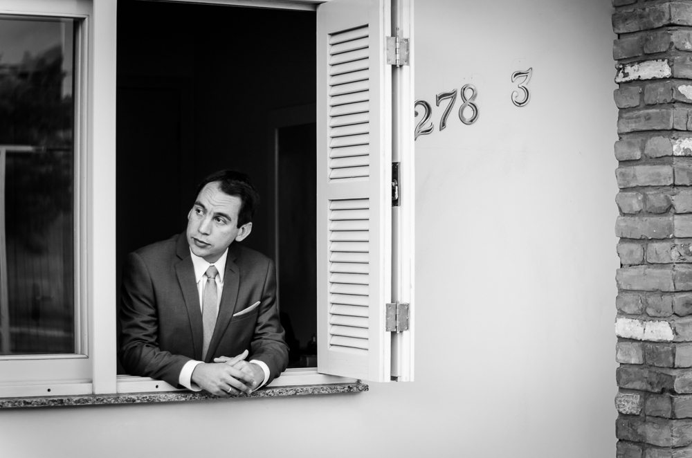 Foto do ensaio de making of do noivo no quarto fazendo os preparativos para o casamento. O noivo já pronto de terno, gravata, lenço e broche olha pela janela de sua casa em Restinga Seca, RS 