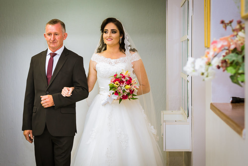 Foto da cerimônia de casamento no Salão do Reino das Testemunhas de Jeová, JW, em Santa Maria, RS. Entrada da noiva de vestido de noiva branco longo, véu e bouquet, buquê, de flores naturais, entra com o pai da noiva de terno e gravata.