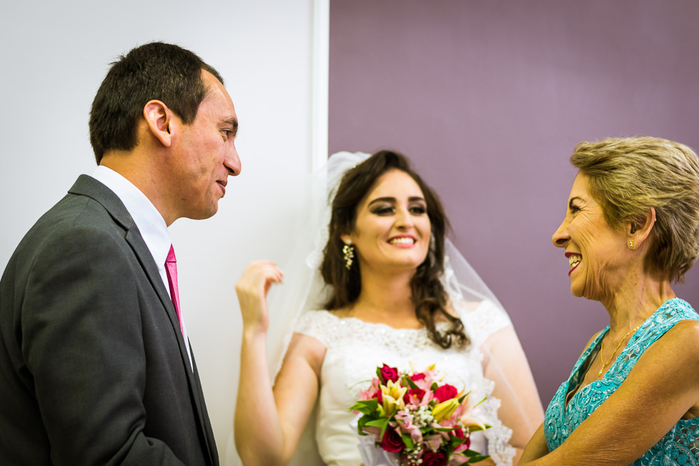 Foto da cerimônia de casamento no Salão do Reino das Testemunhas de Jeová, JW, em Santa Maria, RS. A tia do noivo sorri feliz e cumprimenta o noiva com a noiva ao fundo sorrindo.
