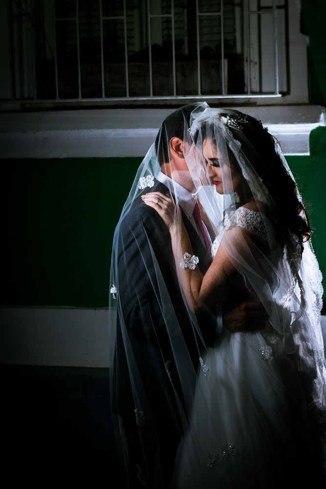 Foto do ensaio fotográfico no dia do casamento do casamento de noivos. Ele de terno,, gravata e ela de vestido de noiva branco, ambos cobertos pelo véu se abraçando na Vila Belga em Santa Maria, RS