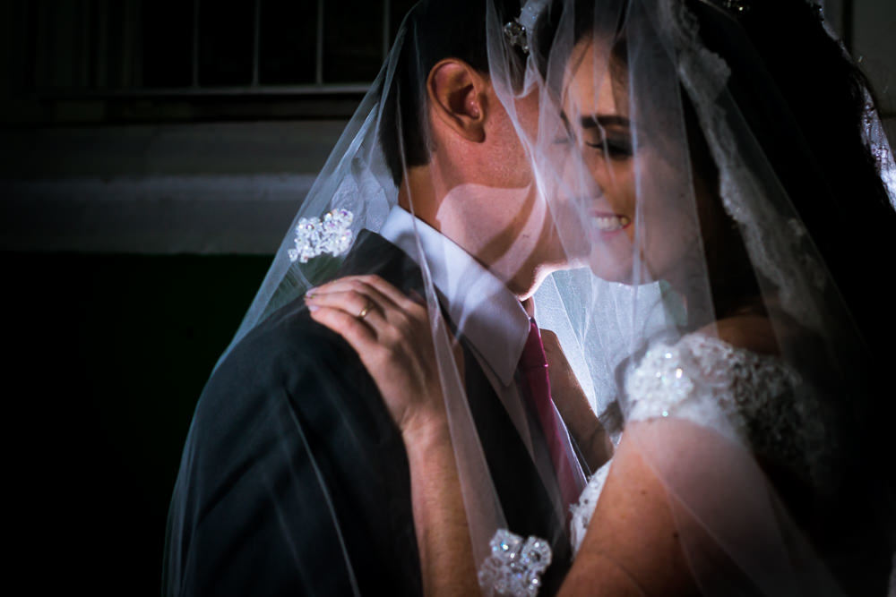 Foto do retrato no ensaio fotográfico no dia do casamento do casamento de noivos. Ele de terno,, gravata e ela de vestido de noiva branco, ambos cobertos pelo véu se abraçando na Vila Belga em Santa Maria, RS