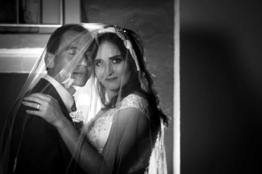 Foto do retrato no ensaio fotográfico no dia do casamento do casamento de noivos. Ele de terno,, gravata e ela de vestido de noiva branco, ambos cobertos pelo véu se abraçando na Vila Belga em Santa Maria, RS