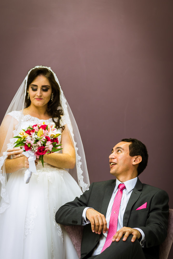 Foto da cerimônia de casamento no Salão do Reino das Testemunhas de Jeová, JW, em Santa Maria, RS. Retrato oficial posada dos noivos, noiva e noivo, ele sentado na cadeira e ela em pé olhando para bouquet, buquê.