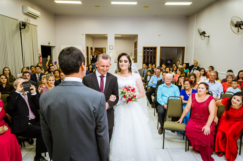 Foto da cerimônia de casamento no Salão do Reino das Testemunhas de Jeová, JW, em Santa Maria, RS. Entrada da noiva de vestido de noiva branco longo, véu e bouquet, buquê, de flores naturais, entra com o pai. O Noivo e convidados olham com alegria.