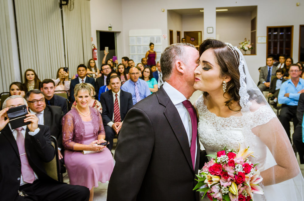 Foto da cerimônia de casamento no Salão do Reino das Testemunhas de Jeová, JW, em Santa Maria, RS. Entrada da noiva de vestido de noiva branco longo, véu e bouquet, buquê, de flores naturais, entra com o pai. O pai da noiva a beija, dá um beijo na filha.
