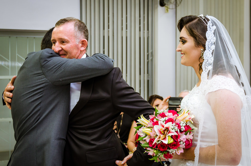 Foto da cerimônia de casamento no Salão do Reino das Testemunhas de Jeová, JW, em Santa Maria, RS. Entrada da noiva de vestido de noiva branco longo, véu e bouquet, buquê, de flores naturais, entra com o pai. O pai da noiva abraça o noivo.