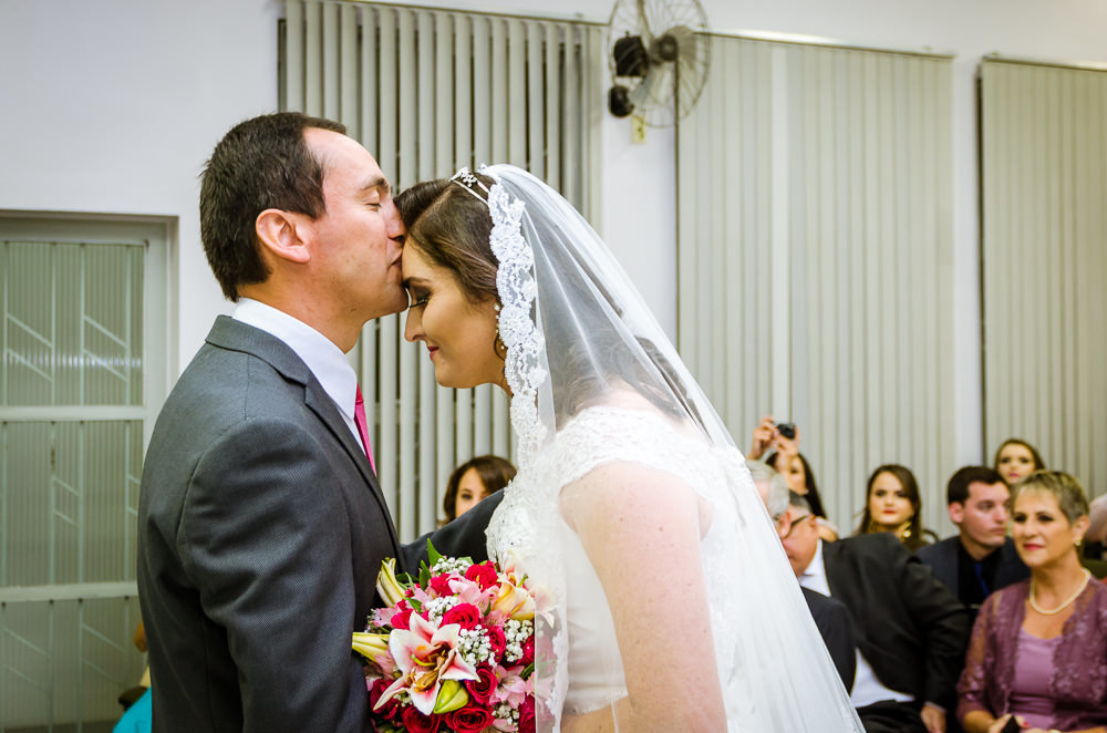 Foto da cerimônia de casamento no Salão do Reino das Testemunhas de Jeová, JW, em Santa Maria, RS. Entrada da noiva de vestido de noiva branco longo, véu e bouquet, buquê, de flores naturais. O noivo beija, dá um beijo na testa da noiva.