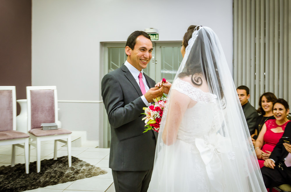 Foto da cerimônia de casamento no Salão do Reino das Testemunhas de Jeová, JW, em Santa Maria, RS. Entrada da noiva de vestido de noiva branco longo, véu e bouquet, buquê, de flores naturais. O noivo surpreende por dar uma flor vermelha para a noiva.
