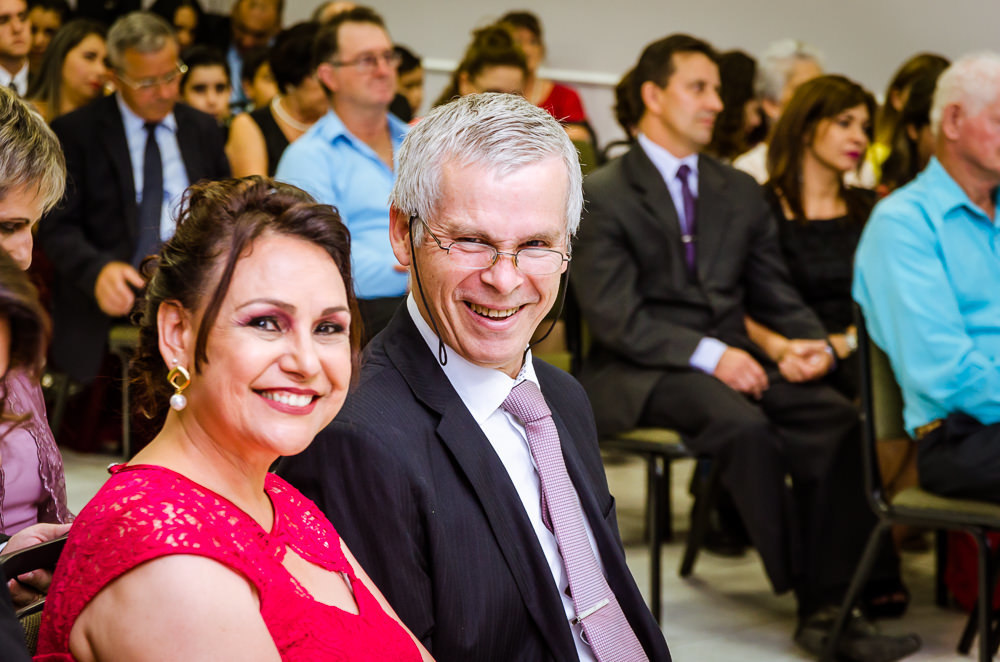 Foto da cerimônia de casamento no Salão do Reino das Testemunhas de Jeová, JW, em Santa Maria, RS. Convidados ao fundo e pai e mãe do noivo estão sorrindo com alegria.