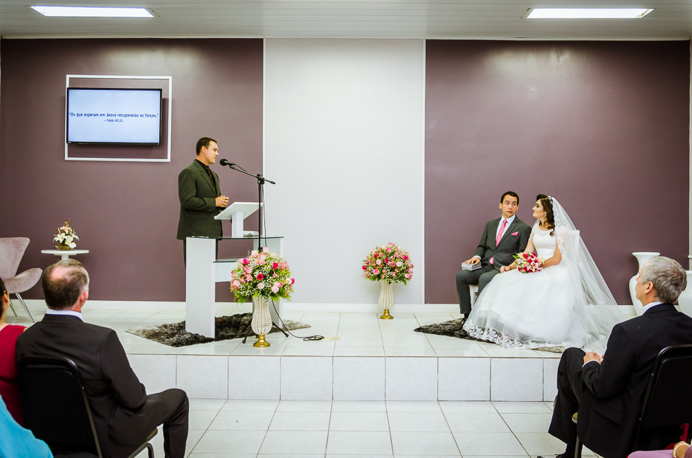Foto da cerimônia de casamento no Salão do Reino das Testemunhas de Jeová, JW, em Santa Maria, RS. Orador do discurso de casamento, noivos sentados se olhando e convidados no auditório.