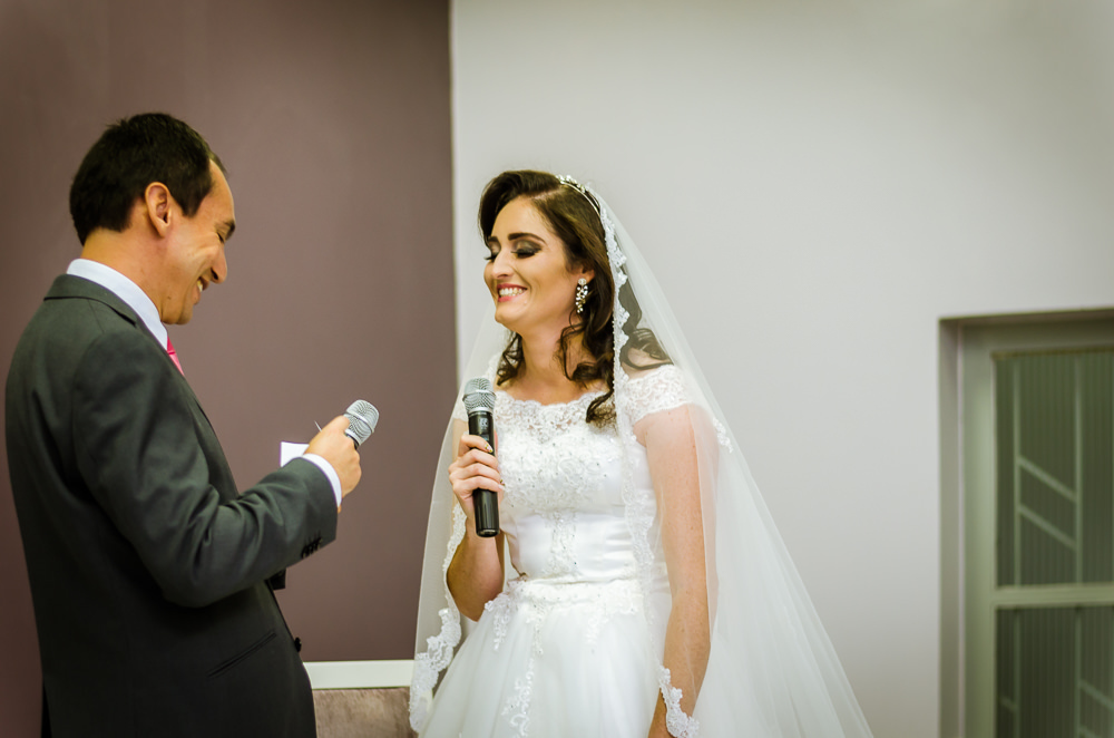 Foto da cerimônia de casamento no Salão do Reino das Testemunhas de Jeová, JW, em Santa Maria, RS. Noivo ouve e ajuda nos votos de casamento da noiva e sorri com ternura.