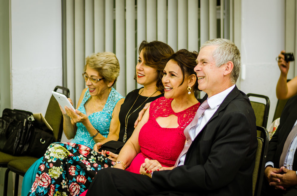 Foto da cerimônia de casamento no Salão do Reino das Testemunhas de Jeová, JW, em Santa Maria, RS. Família da noiva, pai, mãe, tia e cunhada junto aos convidados sorriem.