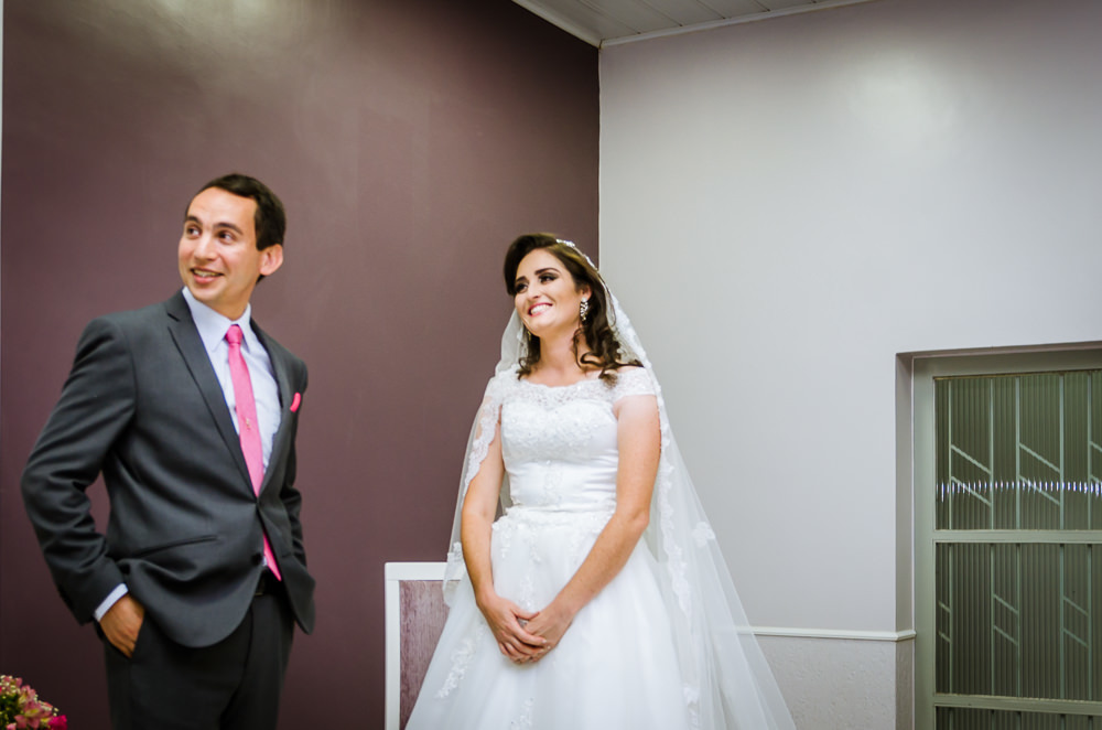 Foto da cerimônia de casamento no Salão do Reino das Testemunhas de Jeová, JW, em Santa Maria, RS. Noivo com as mãos no bolso esquece as alianças de casamento, a noiva sorri.