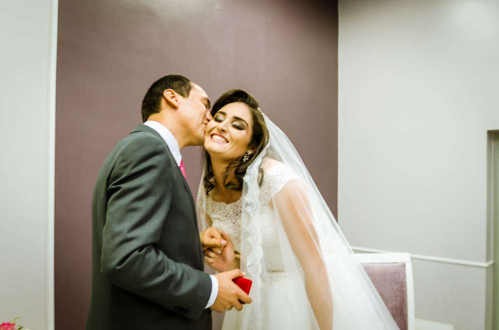 Foto da cerimônia de casamento no Salão do Reino das Testemunhas de Jeová, JW, em Santa Maria, RS. Após a troca das alianças, o noivo beija com carinho a noiva que sorri.