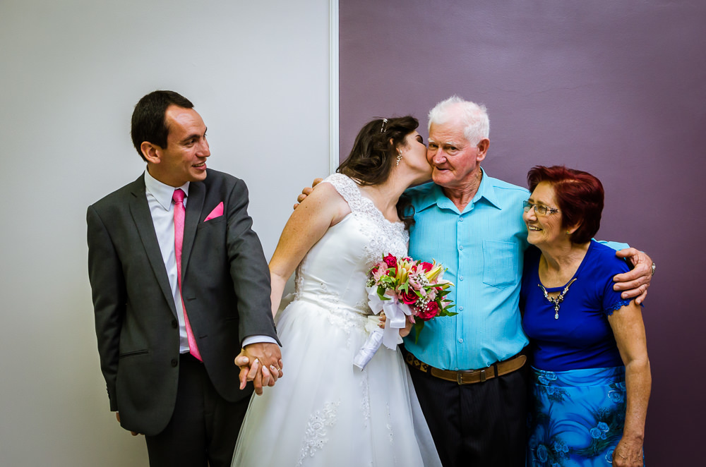 Foto da cerimônia de casamento no Salão do Reino das Testemunhas de Jeová, JW, em Santa Maria, RS. Retrato oficial posada dos noivos com a família da noiva, avô e avó da noiva. A noiva beija com ternura seu avô.