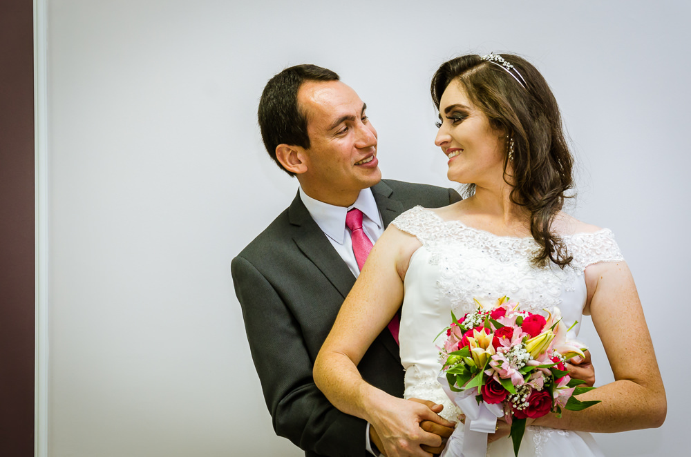 Foto da cerimônia de casamento no Salão do Reino das Testemunhas de Jeová, JW, em Santa Maria, RS. Retrato oficial posada dos noivos, noiva e noivo, ele sentado na cadeira e ela em pé olhando para bouquet, buquê.