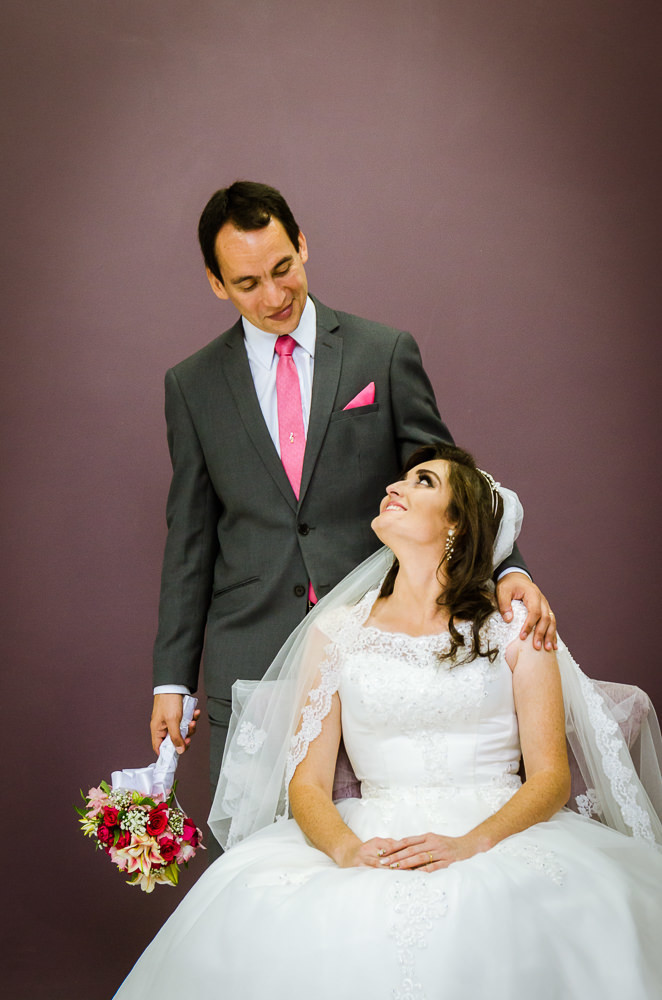 Foto da cerimônia de casamento no Salão do Reino das Testemunhas de Jeová, JW, em Santa Maria, RS. Retrato oficial posada dos noivos, noiva e noivo, ele em pé segura o bouquet, buquê olhando a noiva com ternura.