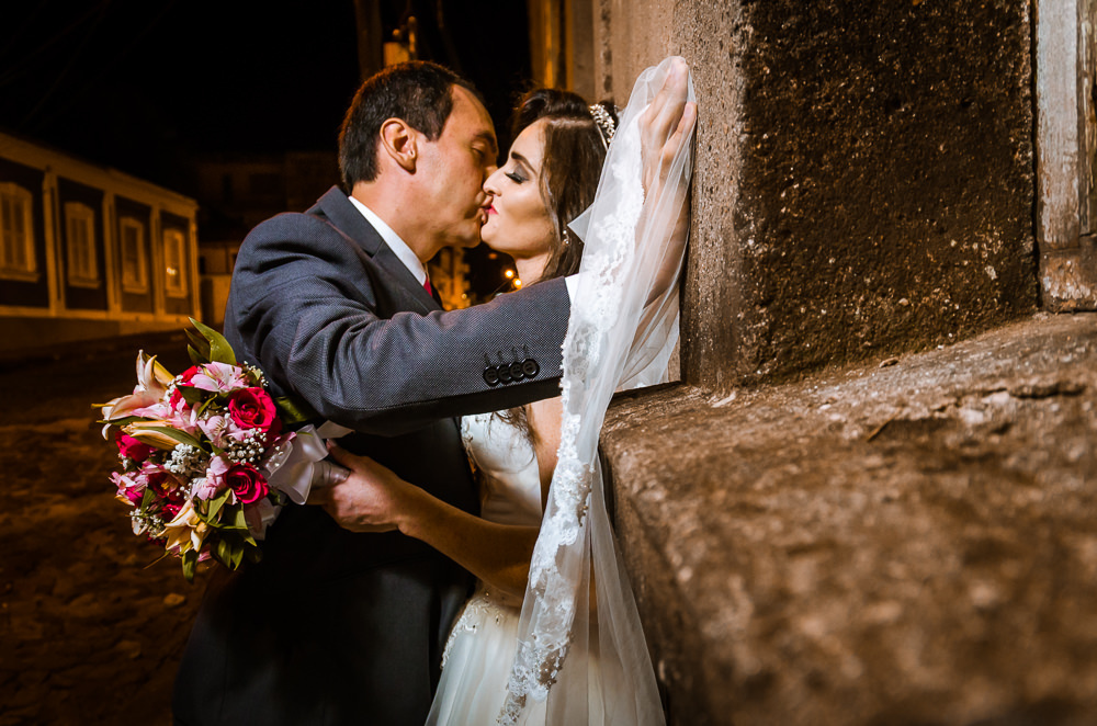 Foto do retrato no ensaio fotográfico no dia do casamento de noivos. Ele de terno, gravata e ela de vestido de noiva branco se beijam, na Vila Belga em Santa Maria, RS