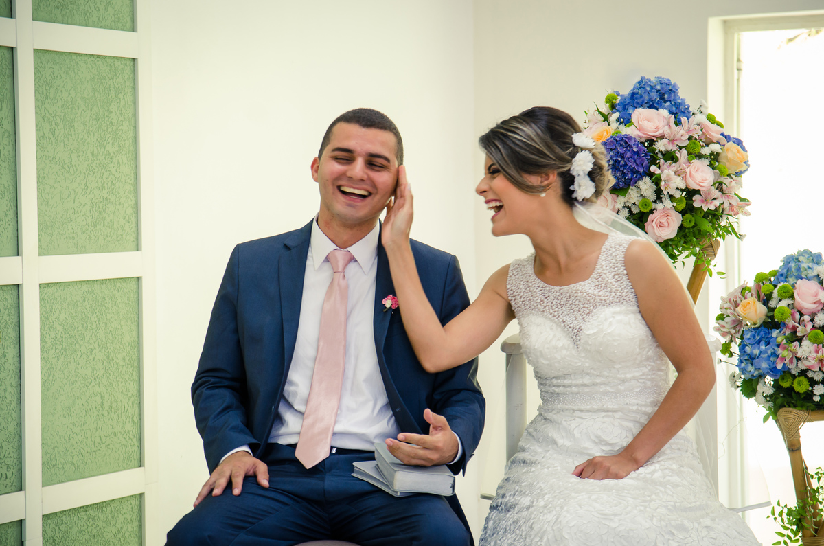 Fotografia do noivo e da noiva sentados sorrindo durante a cerimônia de casamento em Cajamar, São Paulo. O noive de terno azul e gravata rosa, a noiva de vestido branco e véu.