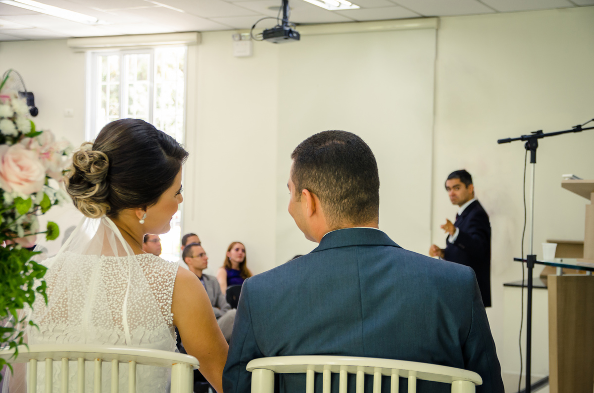 Fotografia do noivo e da noiva sentados sorrindo durante a cerimônia de casamento em Cajamar, São Paulo. O noive de terno azul e gravata rosa, a noiva de vestido branco e véu.