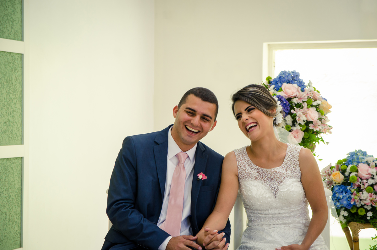 Fotografia do noivo e da noiva sentados sorrindo durante a cerimônia de casamento em Cajamar, São Paulo. O noive de terno azul e gravata rosa, a noiva de vestido branco e véu.
