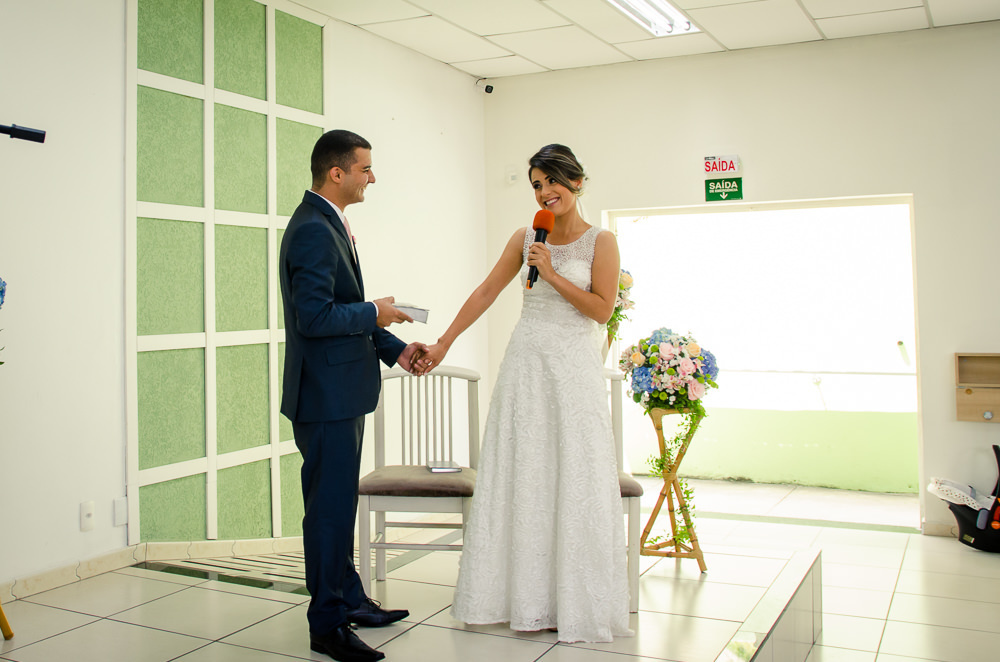 Fotografia do noivo e da noiva sentados sorrindo durante a cerimônia de casamento em Cajamar, São Paulo. O noive de terno azul e gravata rosa, a noiva de vestido branco e véu.