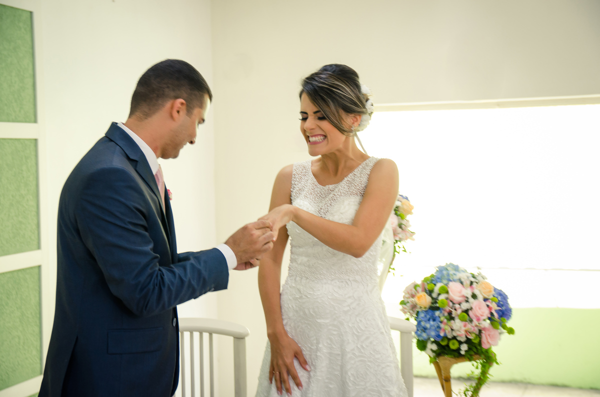 Fotografia do noivo e da noiva sentados sorrindo durante a cerimônia de casamento em Cajamar, São Paulo. O noive de terno azul e gravata rosa, a noiva de vestido branco e véu.