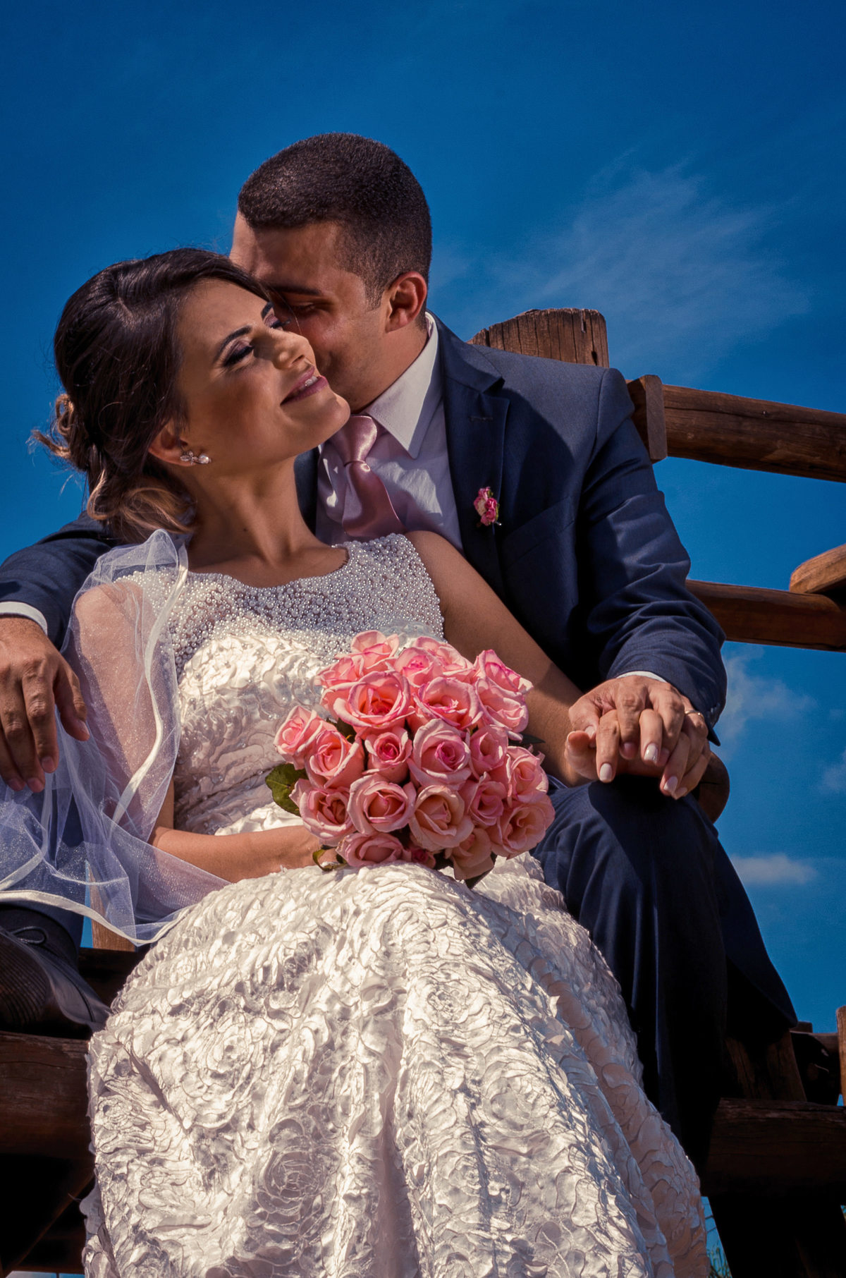 Foto do ensaio após a cerimônia de casamento dos noivos. A noiva está de vestido de noiva branco e véu com o bouquet, buquê, e o noivo de terno azul e gravata rosa em Cajamar, São Paulo