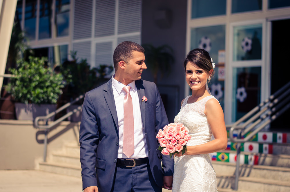 Foto dos noivos chegando a recepção por adesão no Anhanguera Parque Shopping, a noiva está de vestido brando e véu com o bouquet, buquê e o noivo de terno azul e gravata rosa em Cajamar, São Paulo