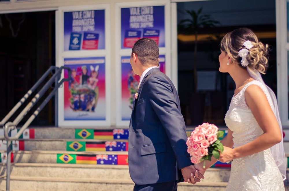 Foto dos noivos chegando a recepção por adesão no Anhanguera Parque Shopping, a noiva está de vestido brando e véu com o bouquet, buquê e o noivo de terno azul e gravata rosa em Cajamar, São Paulo