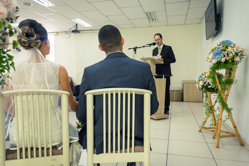 Fotografia do noivo e da noiva sentados sorrindo durante a cerimônia de casamento em Cajamar, São Paulo. O noive de terno azul e gravata rosa, a noiva de vestido branco e véu.
