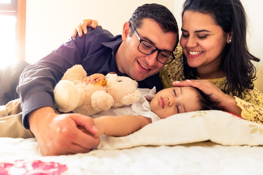 Fotografia Documental de Família. Foto da rotina da família com seu filho criança bebê, mostrando a essência a história da família.  Foto em Barueri, SP. Filho dormindo com ursinho, urso e pais, papai e mamãe sorrindo e admirando seu filho. 