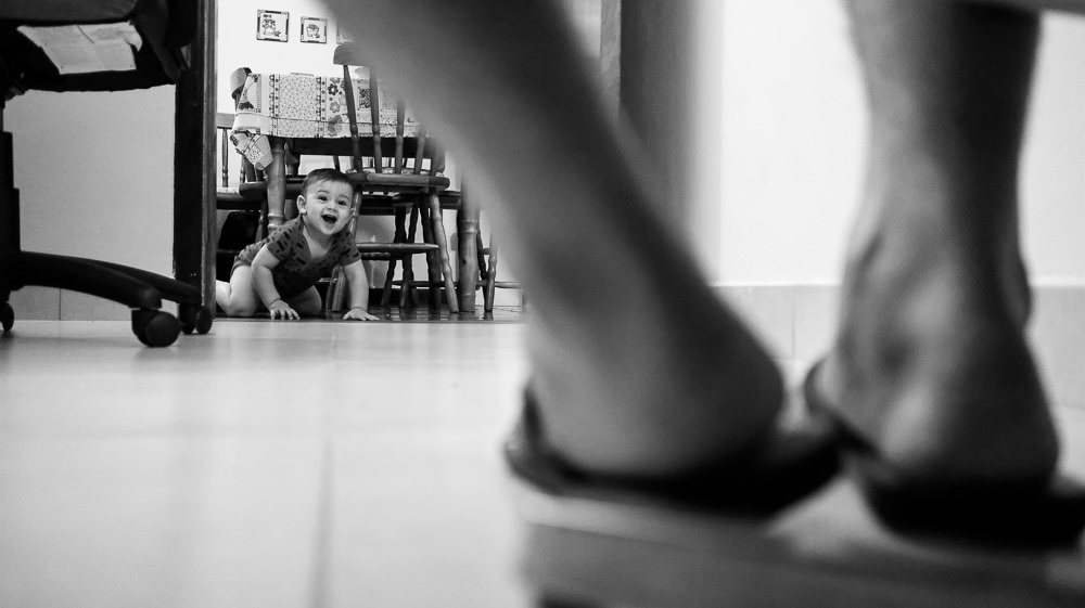 Fotografia documental de Família, Filho bebê engatinhando e sorrindo feliz por ver o papai, pai que está sentado. A foto tem a visão do chão por traz dos pés do pai que está sentado. Em Barueri, SP