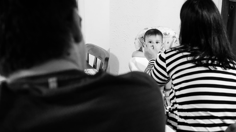 Fotografia documental de Família, Filho bebê, criança na cadeirinha comendo enquanto a mãe, mamãe alimenta, dá papinha, comida na boca, pai olha admirado. Em Barueri, SP