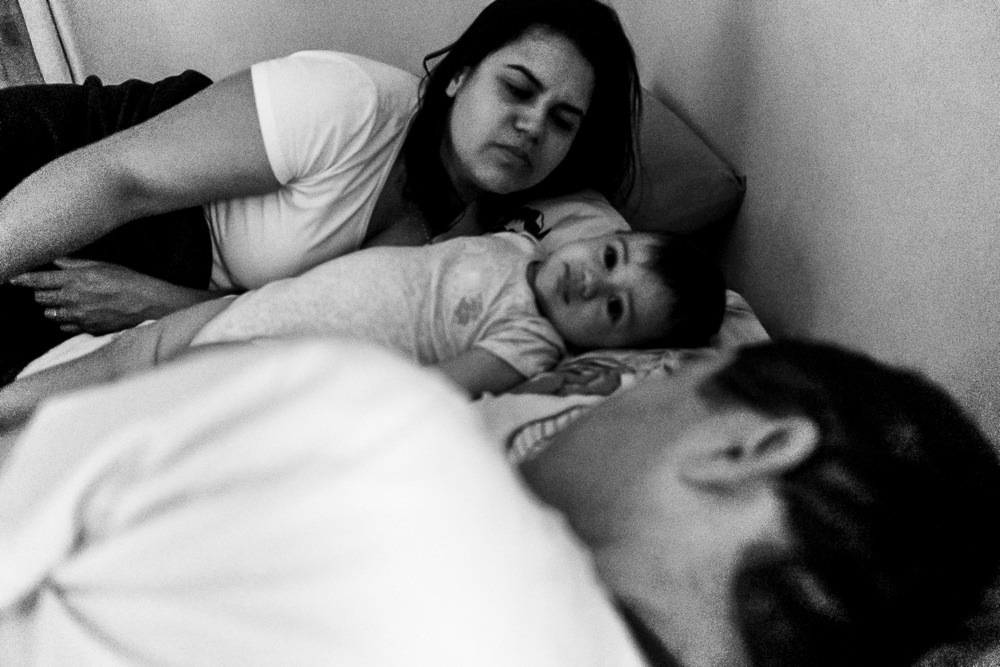 Fotografia documental de Família, Hora de acordar, pai, mãe e filho criança na cama acordando. Foto em PB preto e branco em Barueri SP
