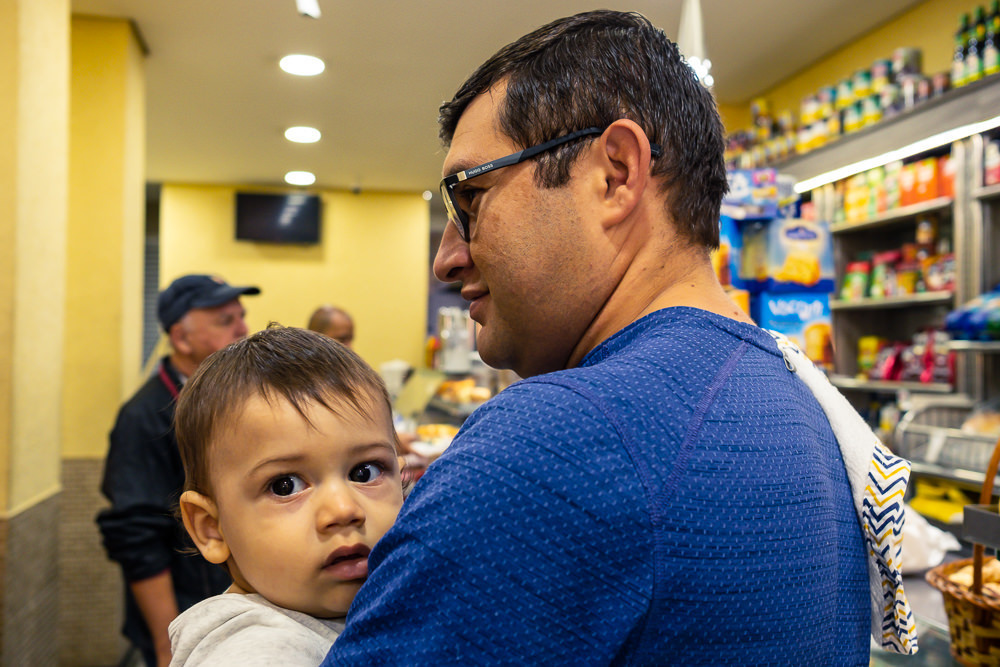 Fotografia Documental de Família. Pai com o filho criança no colo na padaria pela manhã para comprar pão par ao café da manhã. Foto colorida em Barueri, SP