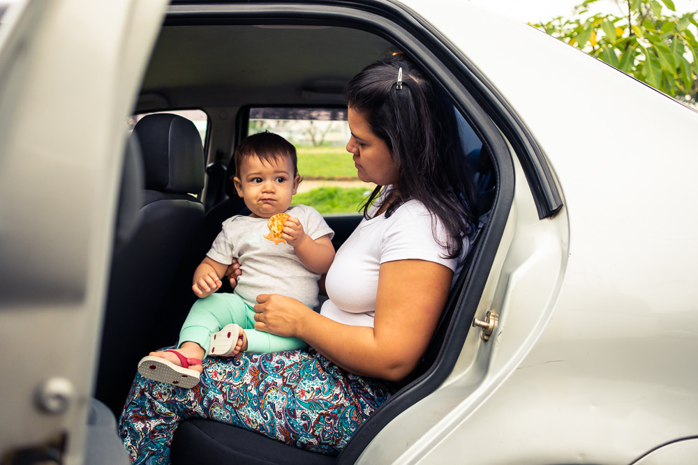 Fotografia Documental de Família, filho criança no colo da mãe, mamãe dentro do carro sentados. A criança está comendo pão de queijo. Foto colorida em Barueri, SP