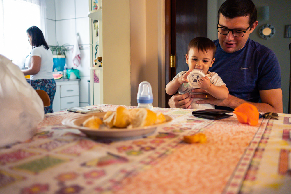 Fotografia Documental de Família. Foto da rotina da família com seu filho criança bebê, mostrando a essência a história da família.  Foto em Barueri, SP. Pai e filho sentados a mesa e mãe ao fundo preparando o café da manhã.