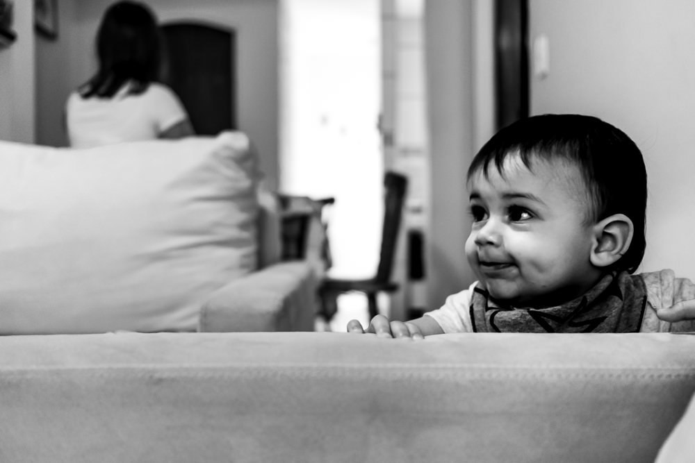 Fotografia Documental de Família. Foto da rotina da família com seu filho criança bebê, mostrando a essência a história da família.  Foto em Barueri, SP. Mãe ao fundo e filho criança, bebê, em pé se apoiando no sofá.