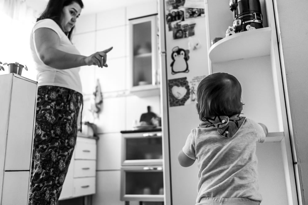 Fotografia Documental de Família. Foto da rotina da família com seu filho criança bebê, mostrando a essência a história da família.  Foto em Barueri, SP. Filho criança em pé se apoiando na estante e mexendo nos objetos como uma criança curiosa.
