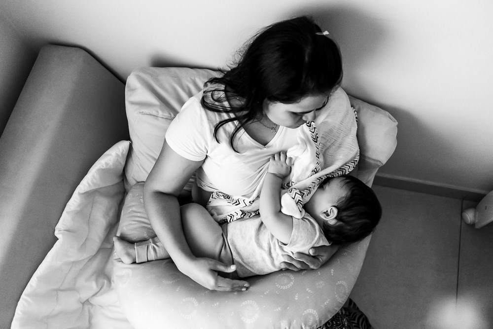 Fotografia Documental de Família. Foto da rotina da família com seu filho criança bebê, mostrando a essência a história da família.  Foto em Barueri, SP. Visto de cima filho criança no colo da mãe mamando no peito, foto em preto e branco pb
