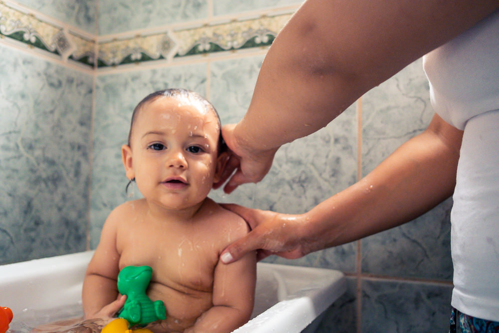 Fotografia Documental de Família. Foto da rotina da família com seu filho criança bebê, mostrando a essência a história da família.  Foto em Barueri, SP. Mamãe dando banho no filho na banheira dentro do box do banheiro com brinquedos