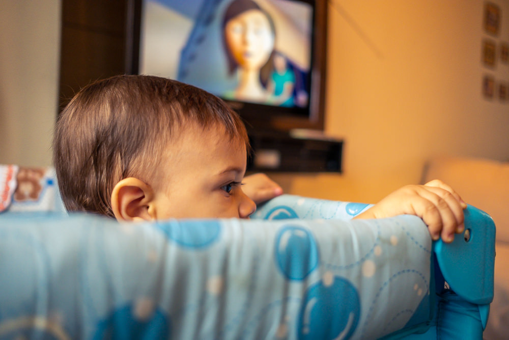 Fotografia Documental de Família. Foto da rotina da família com seu filho criança bebê, mostrando a essência a história da família.  Foto em Barueri, SP. Criança no chiqueirinho cercado com TV ao fundo assistindo video do Pedrinho e Sofia do TV JW 