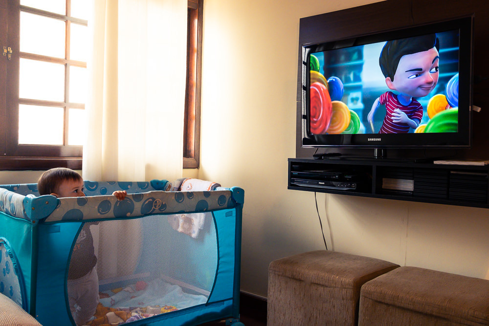 Fotografia Documental de Família. Foto da rotina da família com seu filho criança bebê, mostrando a essência a história da família.  Foto em Barueri, SP. Criança no chiqueirinho cercado com TV ao fundo assistindo video do Pedrinho e Sofia do TV JW 