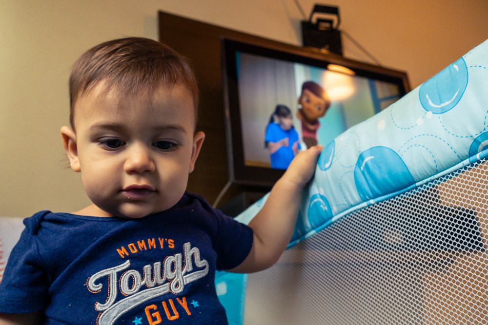 Fotografia Documental de Família. Foto da rotina da família com seu filho criança bebê, mostrando a essência a história da família.  Foto em Barueri, SP. Criança no chiqueirinho cercado com TV ao fundo assistindo video do Pedrinho e Sofia do TV JW 